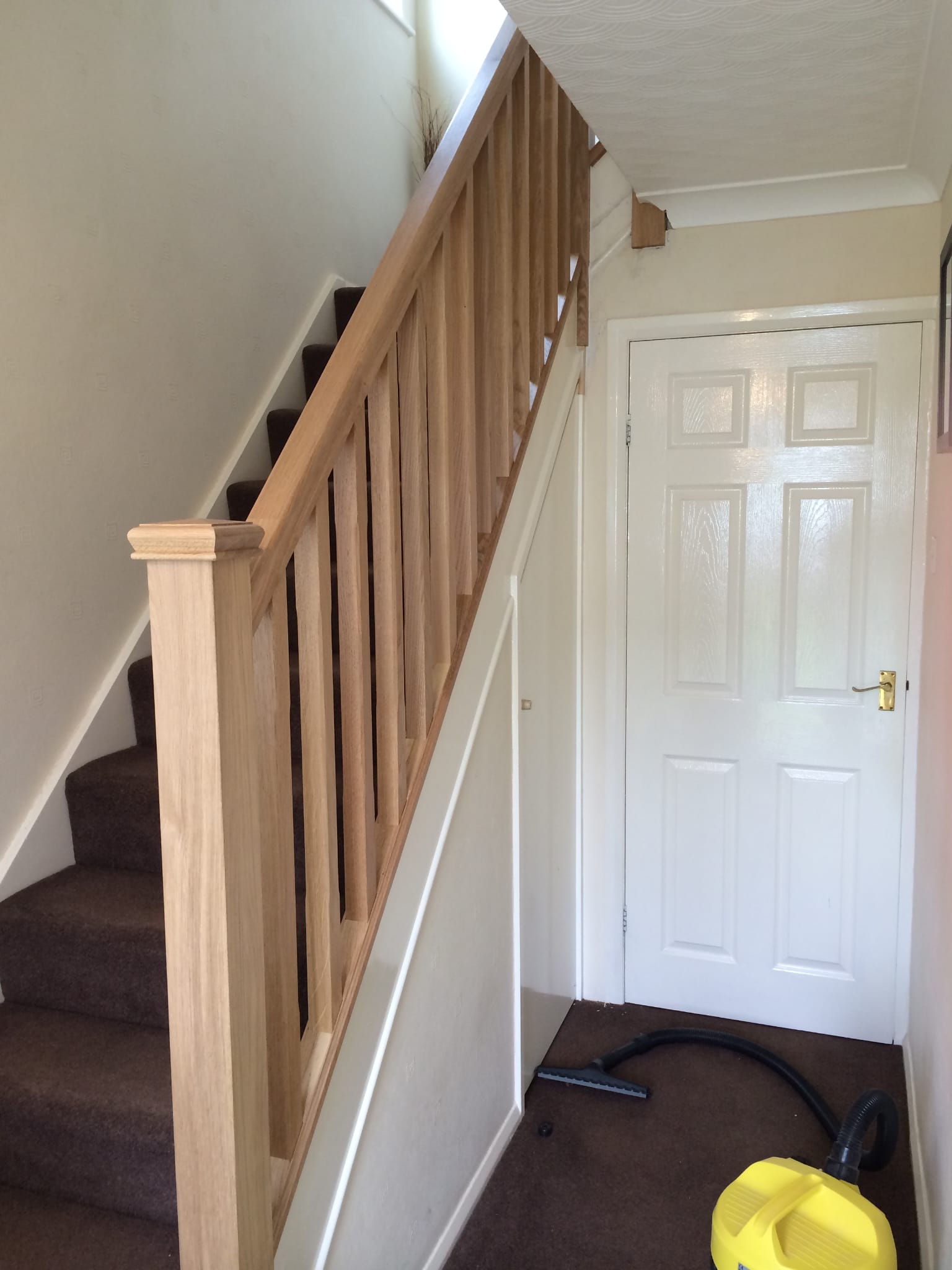 A hallway featuring a white door with a golden handle and a wooden staircase with brown carpeting, showcasing fine joinery. A yellow vacuum cleaner is visible on the brown carpet floor near the base of the stairs. The walls are painted white, and the area is well-lit. - TJS Bathrooms & Kitchens Ltd