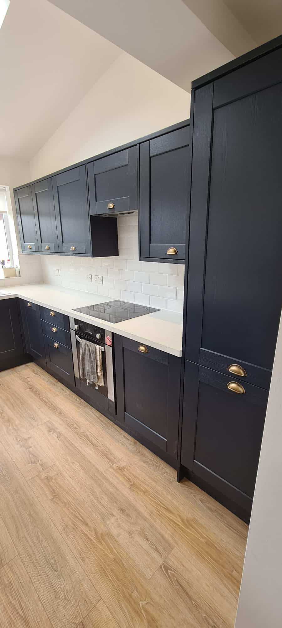 A modern kitchen featuring dark blue shaker-style cabinets with brass handles, a white tile backsplash, and light-colored wooden flooring. The kitchen includes a built-in oven and stove, with a towel hanging on the oven door handle. - TJS Bathrooms & Kitchens Ltd