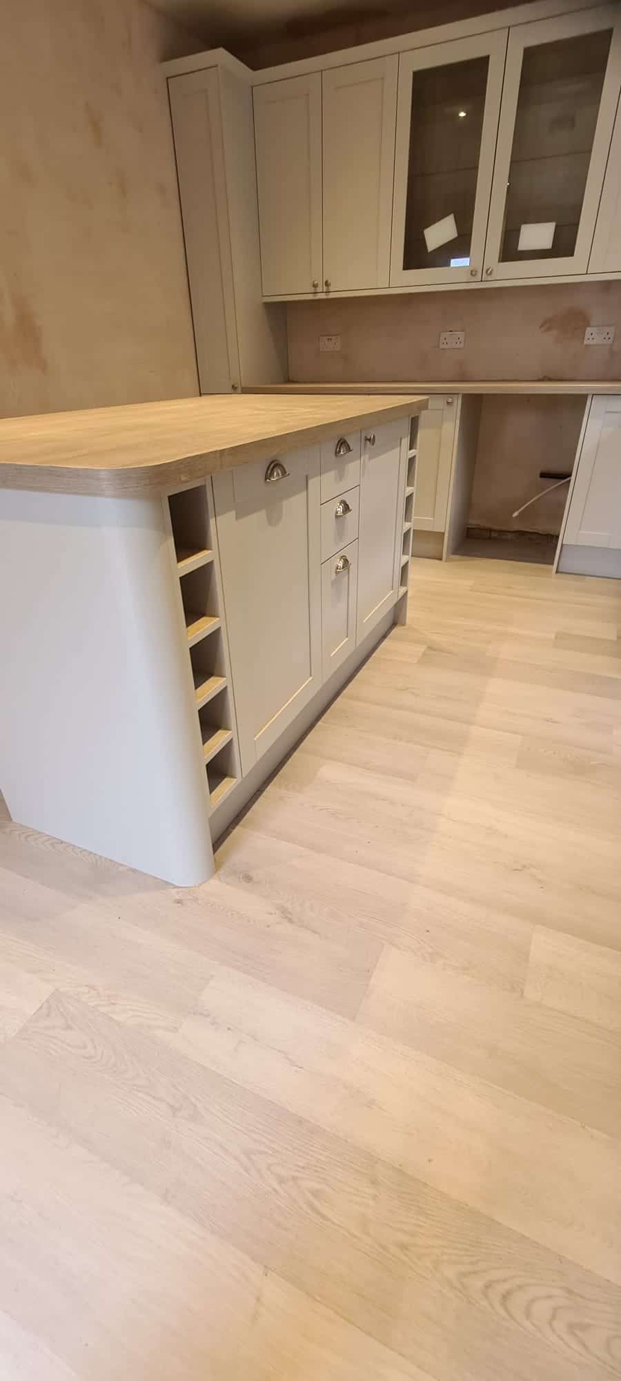 A modern kitchen interior featuring a light wooden floor, light-colored cabinetry with glass-paneled cabinet doors, and a central island with a wood countertop. The island includes built-in shelving and drawers. The overall design is minimalist and clean. - TJS Bathrooms & Kitchens Ltd