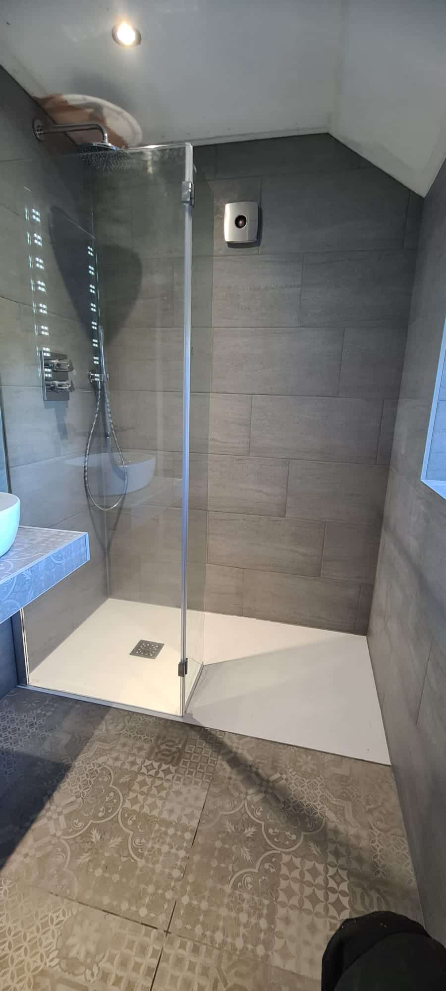 A modern bathroom with a glass-paneled shower. The shower has a chrome rain shower head and gray tiled walls. Adjacent to the shower is a patterned tile floor and a section of the sink countertop with a white vessel sink partially visible. - TJS Bathrooms & Kitchens Ltd