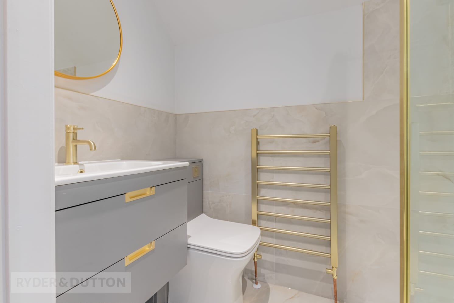 A modern bathroom features a sleek gray vanity with gold fixtures, a white toilet, and a polished brass towel rack. The walls are clad in light, marble-textured tiles, and a frameless glass shower is partially visible. The sink includes a round mirror above it. - TJS Bathrooms & Kitchens Ltd